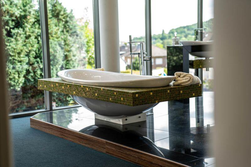 Freistehende Badewanne auf einem Podest mit grünem Glasmosaik vor der Glasfront der Ausstellung Bonn